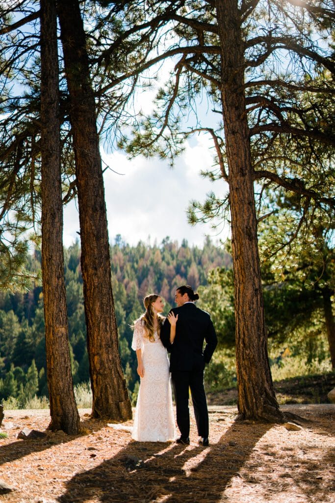A couple dressed in wedding attire stands closely together in a sunlit forest, surrounded by tall pine trees, with green mountains in the background.