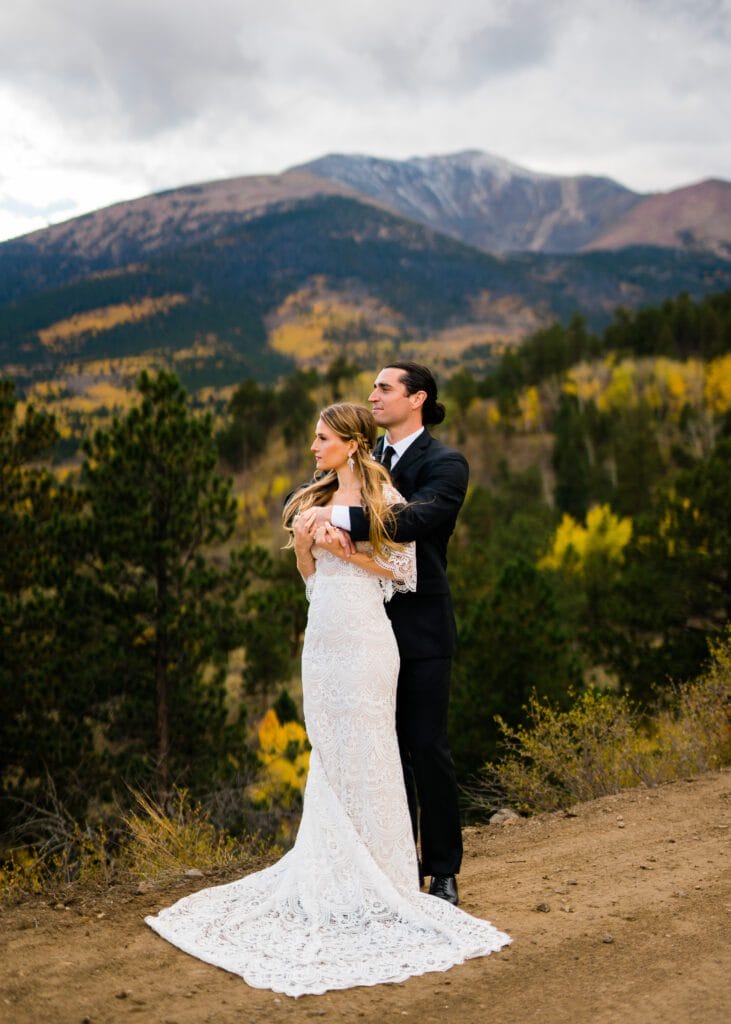 A bride in a white lace gown and a groom in a black suit embrace on a dirt path, surrounded by pine trees, with mountains and cloudy skies in the background.