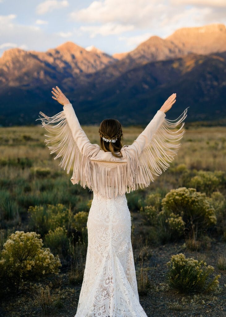 A woman in a white lace dress and a fringed jacket stands in a field with arms raised, facing distant mountains under a partly cloudy sky at sunset.
