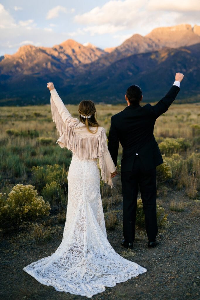 A bride and groom stand hand in hand, raising their arms triumphantly while facing distant mountains in a scenic outdoor setting. The bride wears a lace gown and fringed jacket; the groom is in a black suit.