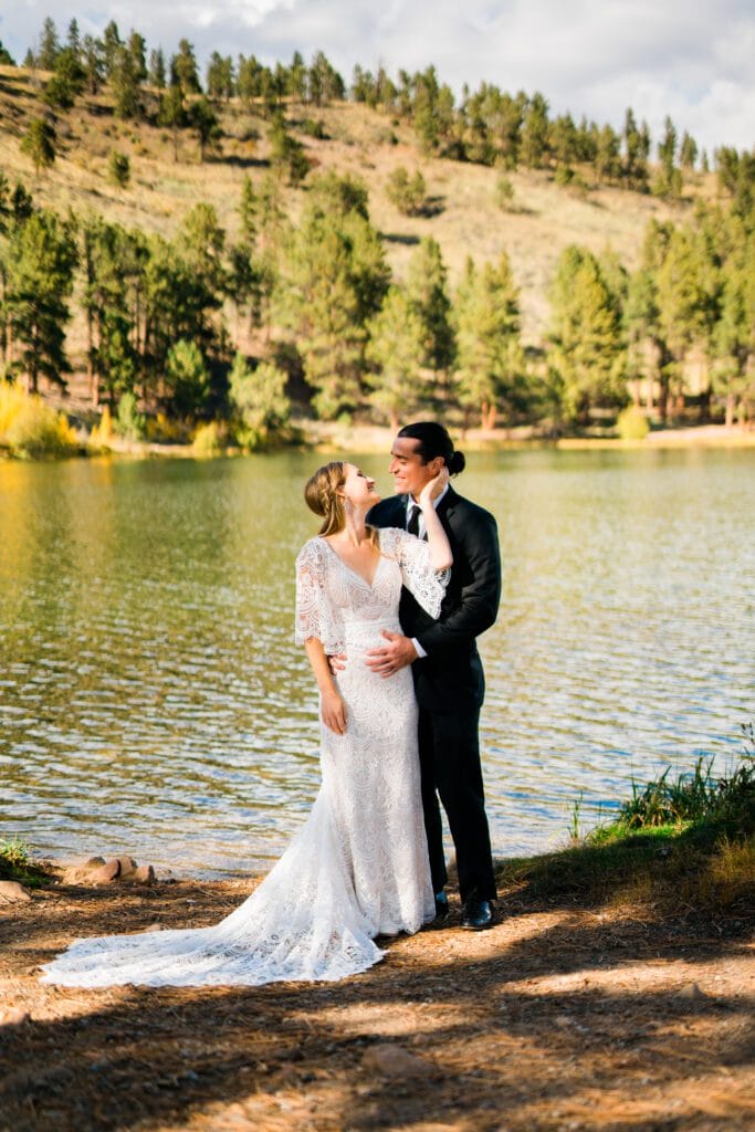 A couple in wedding attire stands by a lake, surrounded by trees and hills. The bride in a white lace gown gazes up at the groom in a black suit, who holds her waist as they smile at each other in the sunlight.