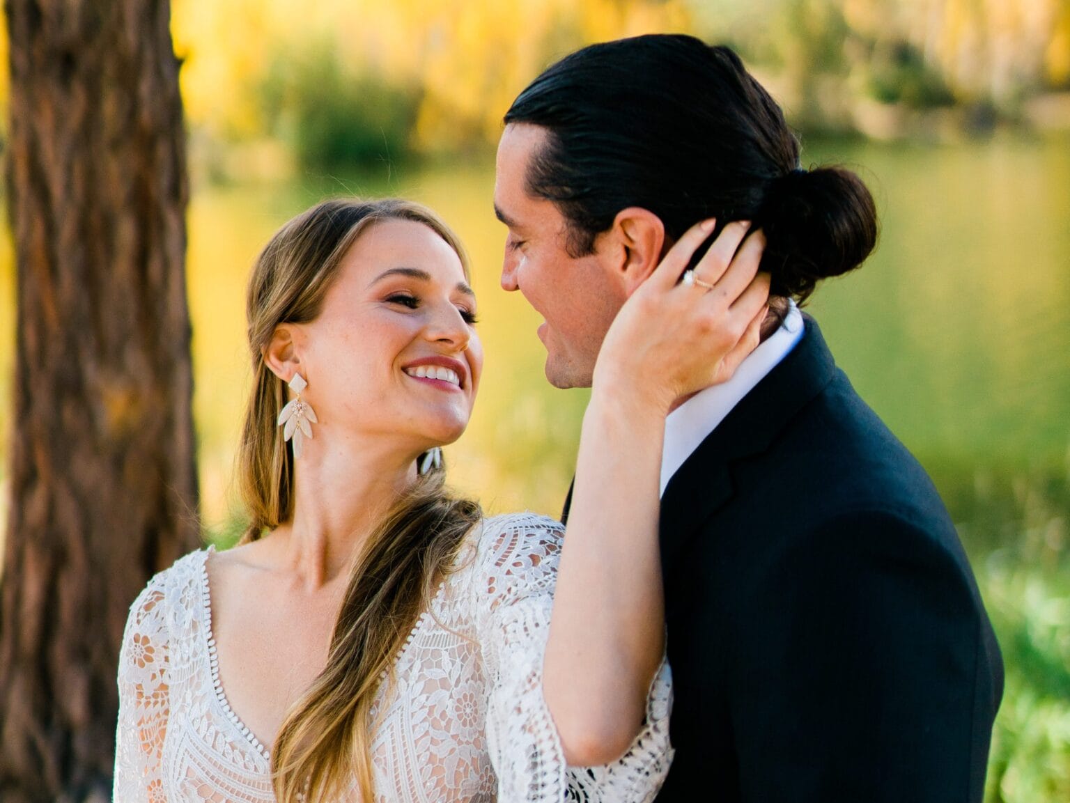 A smiling couple dressed formally embrace outdoors by a lake, with the woman’s hand touching the mans neck and both gazing happily at each other. Green trees and water are blurred in the background.