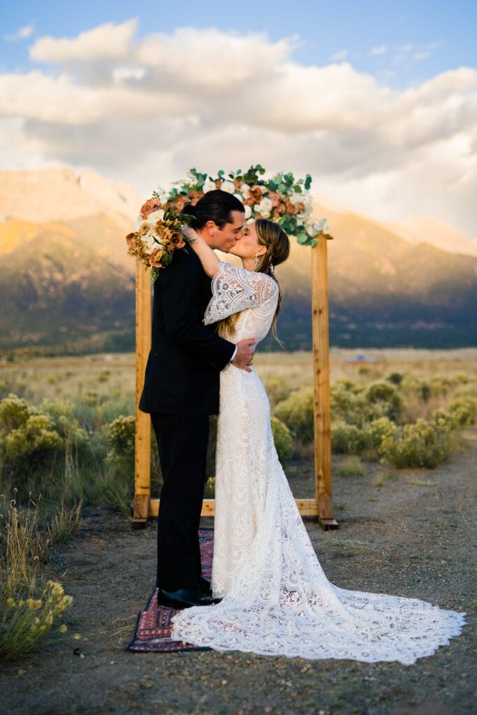 A bride and groom share a kiss outdoors in front of a wooden floral arch, with mountains and cloudy sky in the background. The bride wears a lace dress and the groom is in a black suit.