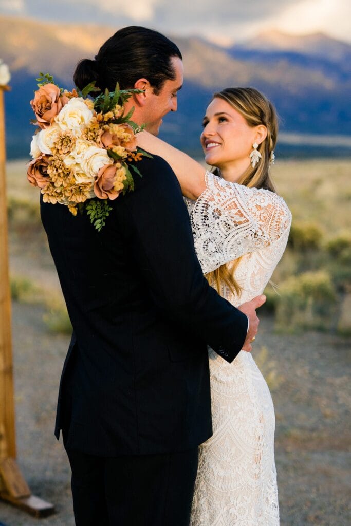A bride in a white lace dress and a groom in a dark suit embrace outdoors, smiling at each other. The bride holds a bouquet of white and brown flowers. Mountains and fields are visible in the background.