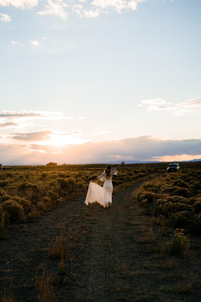 A woman in a white dress walks down a dirt path through a field at sunset, holding her dress out to the sides. The sky is partly cloudy, and a car is parked off the road in the background.