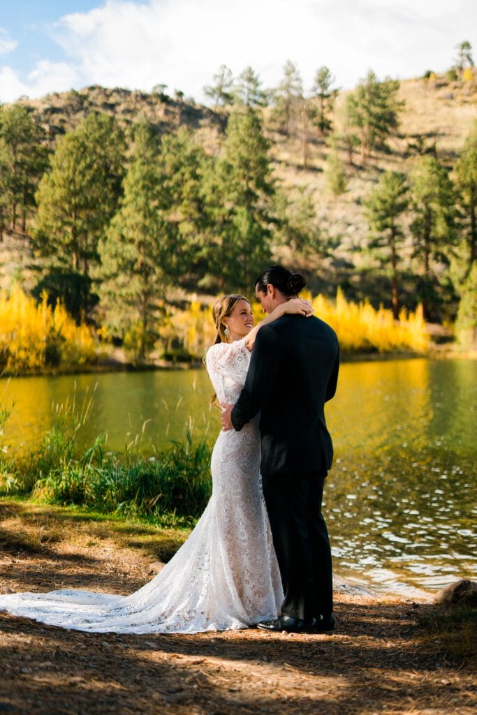 A bride and groom embrace by a peaceful lake surrounded by trees and autumn foliage, with sunlight illuminating the scene and a forested hillside in the background.