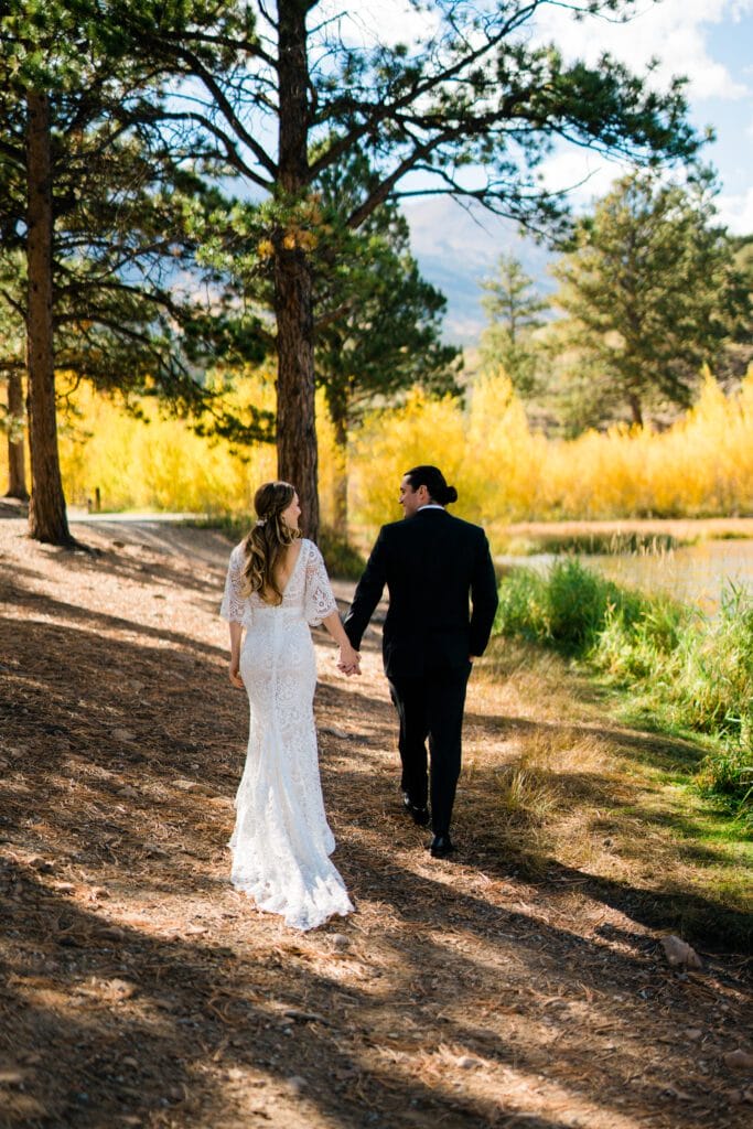A bride and groom walk hand in hand along a sunlit wooded path, surrounded by tall trees and yellow autumn foliage. The bride wears a white lace gown, and the groom is in a black suit.