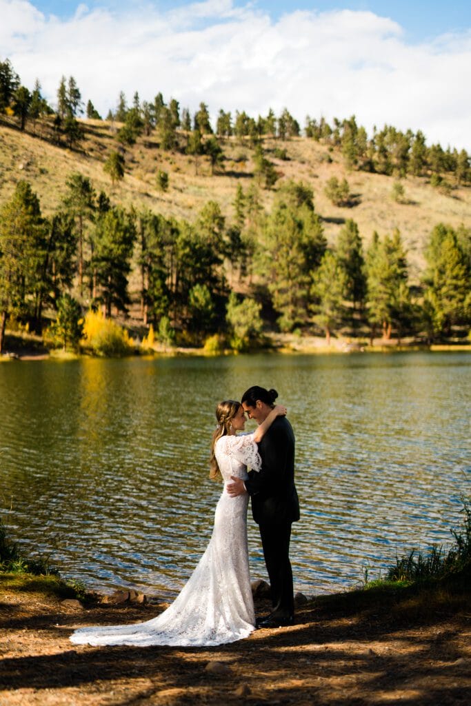 A bride and groom embrace by the edge of a lake, surrounded by pine-covered hills under a bright sky, with sunlight highlighting the bride’s white dress and the tranquil water behind them.