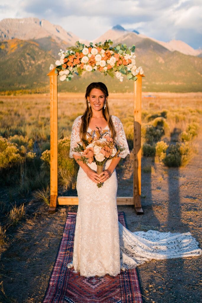 A bride in a white lace gown holds a bouquet and stands on a rug under a floral arch outdoors, with mountains and golden fields in the background, during sunset.