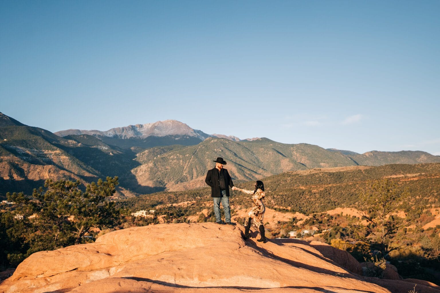 boyfriend helping his girlfriend up a steep rock with pikes peak in the background during their garden of the gods photoshoot