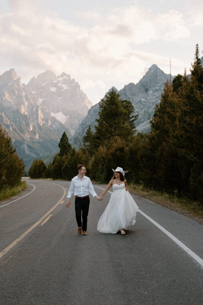 A bride in a white dress and hat walks hand-in-hand with a groom in a white shirt and dark pants on a road, surrounded by trees and mountains under a cloudy sky.