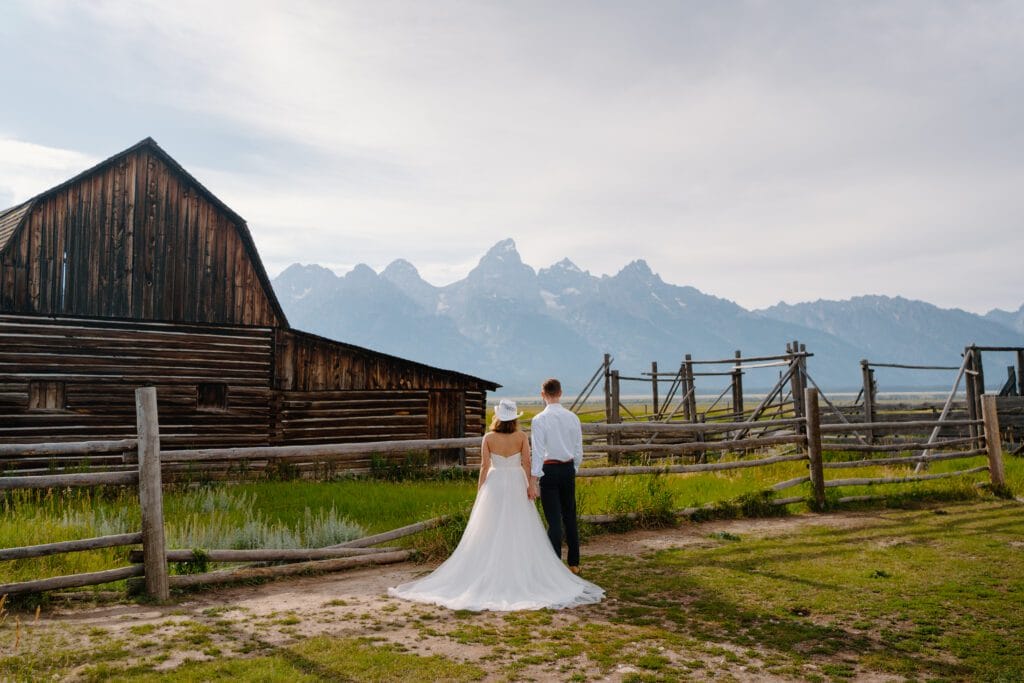A bride and groom stand together facing away, looking at mountains beyond a rustic wooden barn and fence under a cloudy sky. The bride wears a white dress and sunhat; the groom wears dress clothes.
