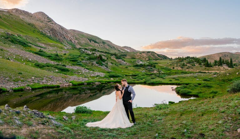 A bride and groom stand by a calm mountain lake surrounded by green hills at sunset, sharing a kiss in their wedding attire with scenic nature in the background.
