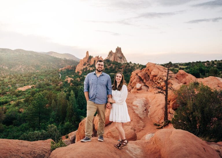 A couple stands on a rocky, red landscape surrounded by greenery and distant rock formations. The man wears a blue shirt and khaki pants; the woman wears a white dress. Both smile at the camera under a bright sky.