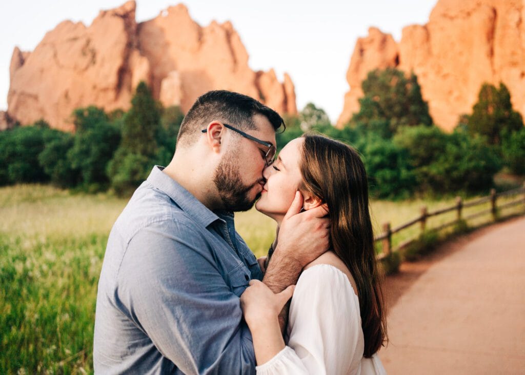 A couple kisses passionately on a path surrounded by greenery, with large reddish rock formations in the background. The man wears glasses and a blue shirt; the woman has long brown hair and a white dress.