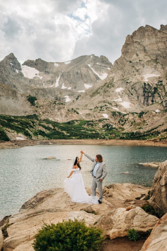 A bride and groom stand on rocky terrain by a mountain lake, surrounded by rugged peaks with patches of snow, as the groom twirls the bride, both smiling under a cloudy sky.