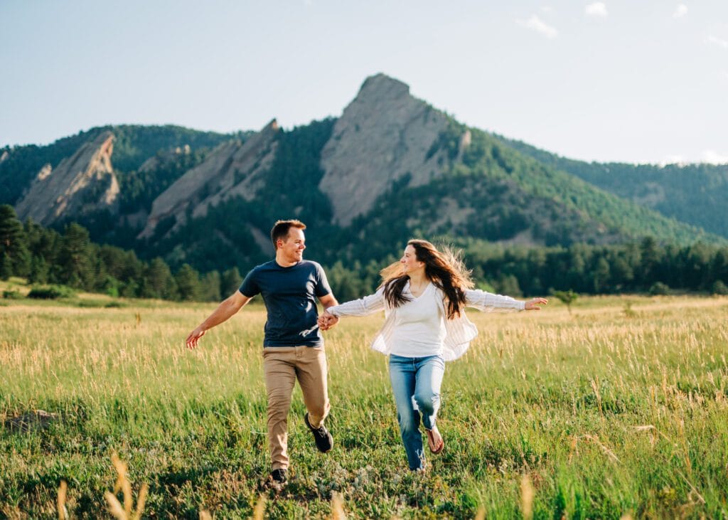 engaged couple holding hands and running towards the camera with the flatiron mountains behind them during their boulder engagement session