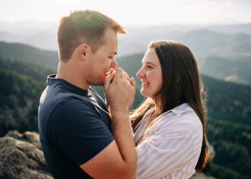 cute engaged couple smiling at each other while fiance grabs girlfriends hands and kisses them during their boulder engagement session