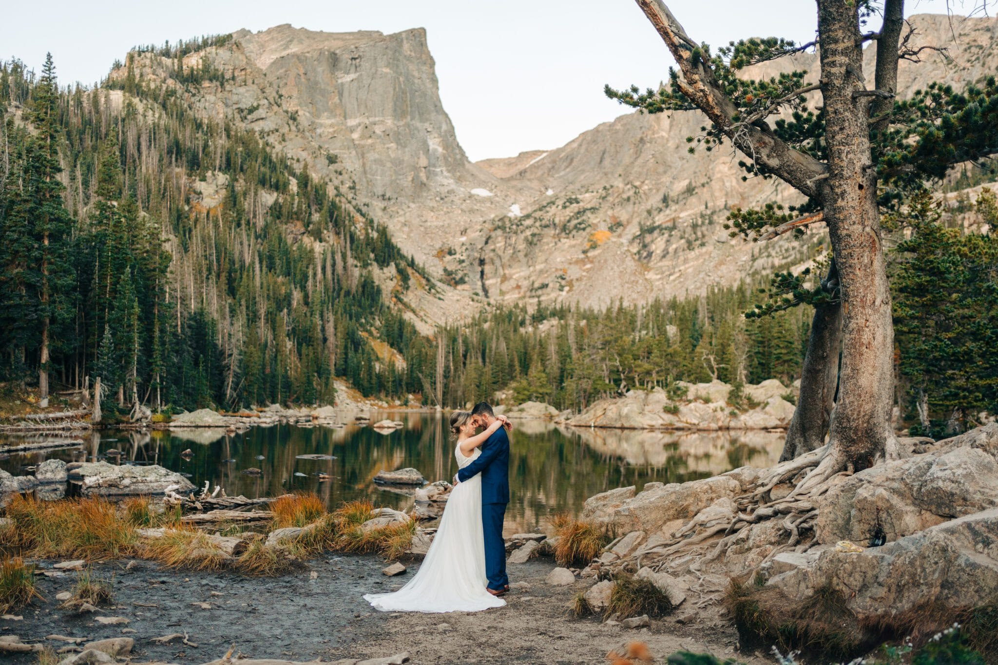 A bride and groom embrace beside a calm mountain lake in Rocky Mountain National Park, surrounded by rocks, trees, and towering cliffs under a clear sky—an unforgettable elopement moment.