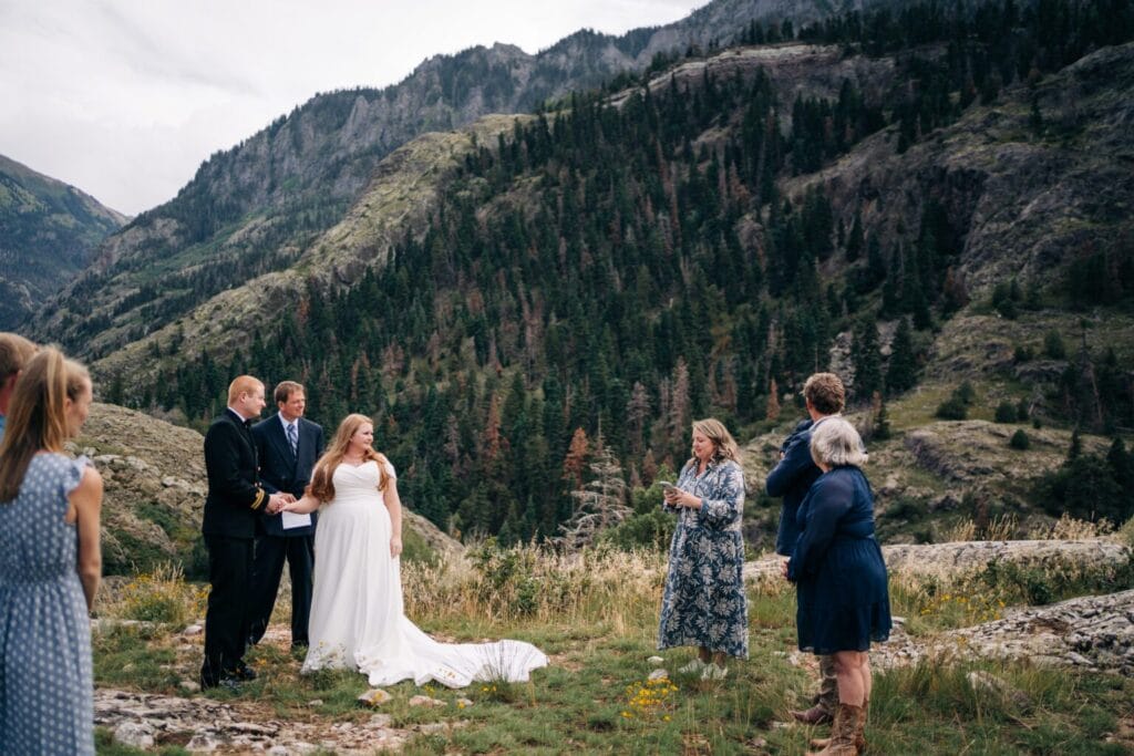 A bride in a white dress stands with a groom and guests in an outdoor wedding ceremony, set against a backdrop of green mountains and pine trees under a cloudy sky.