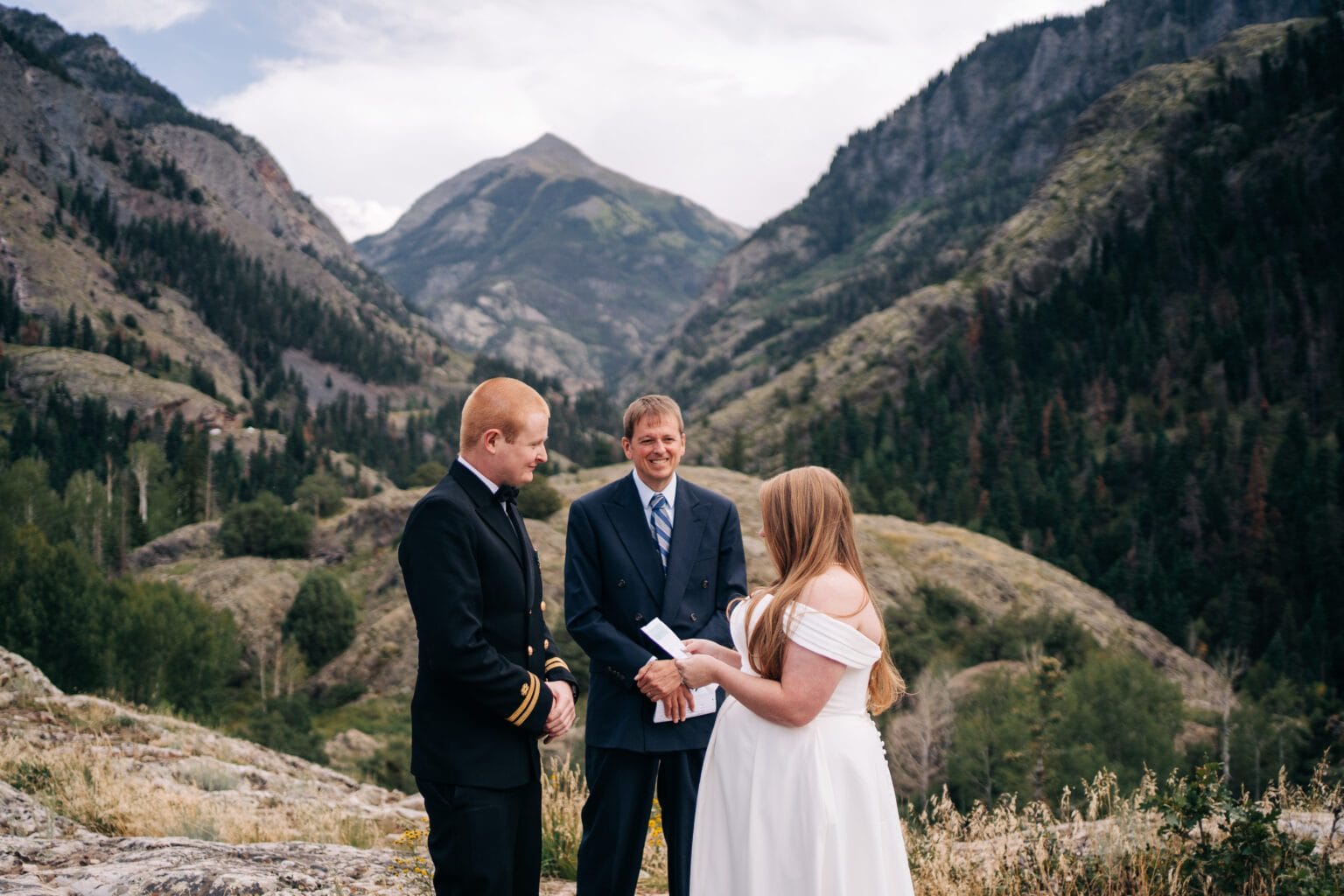 A couple stands with an officiant in formal attire, exchanging vows outdoors in a mountainous landscape with greenery and dramatic peaks in the background.