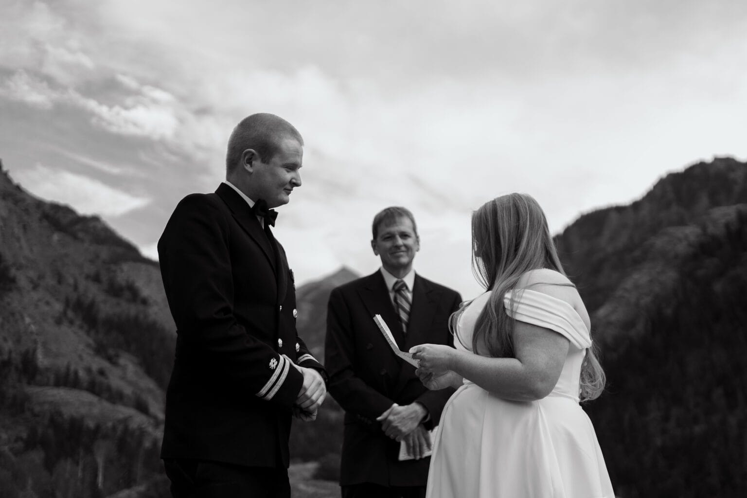 A bride and groom stand outdoors facing each other, with a person officiating between them. Mountains and trees are visible in the background under a cloudy sky. The image is in black and white.