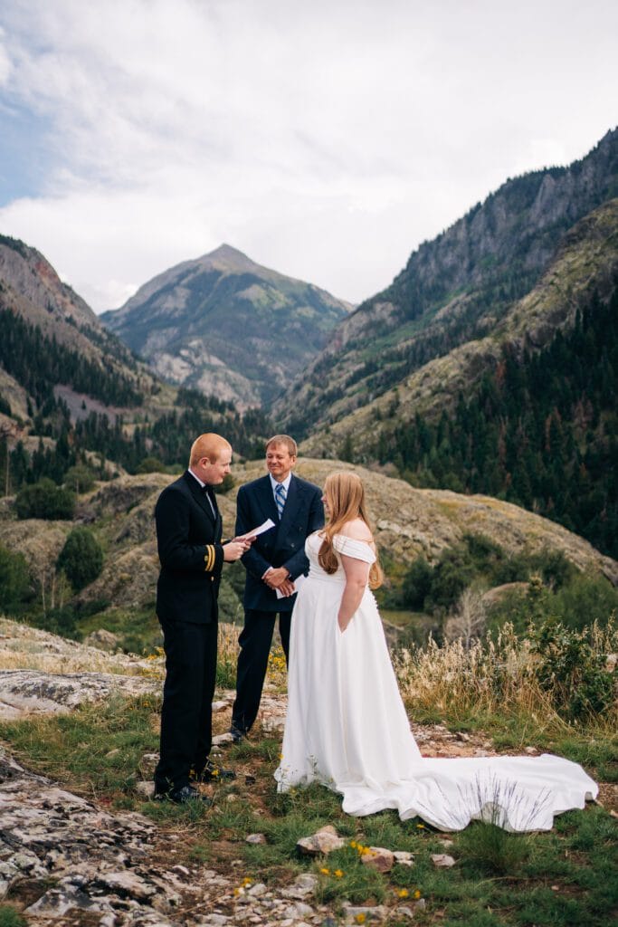 A couple stands with an officiant on a rocky hillside, surrounded by mountains and greenery. The groom wears a black military uniform, and the bride wears a white dress as they exchange vows outdoors.