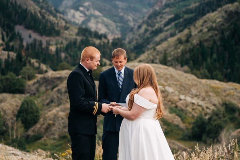 A couple stands exchanging rings during a wedding ceremony outdoors, with a man officiating. They are surrounded by mountains and greenery, creating a scenic backdrop.