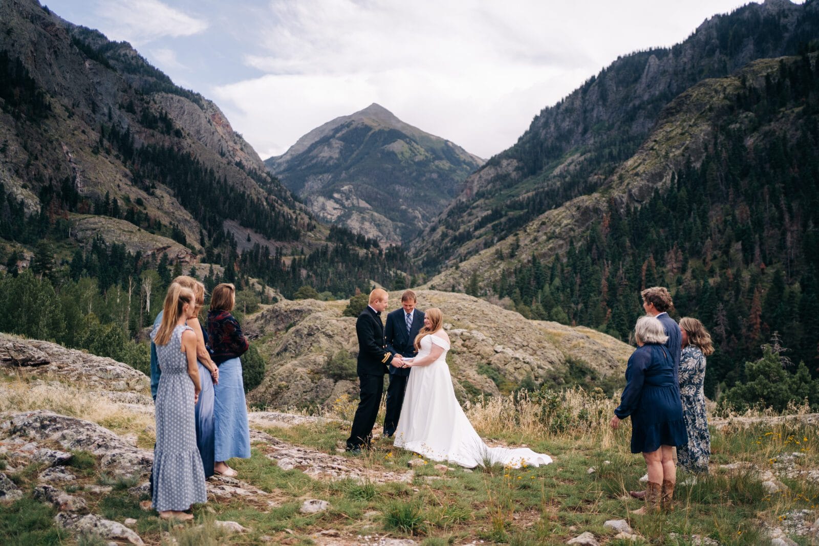 A bride and groom stand holding hands during a wedding ceremony on a grassy mountain slope, surrounded by a small group of guests, with tall mountains and trees in the background.