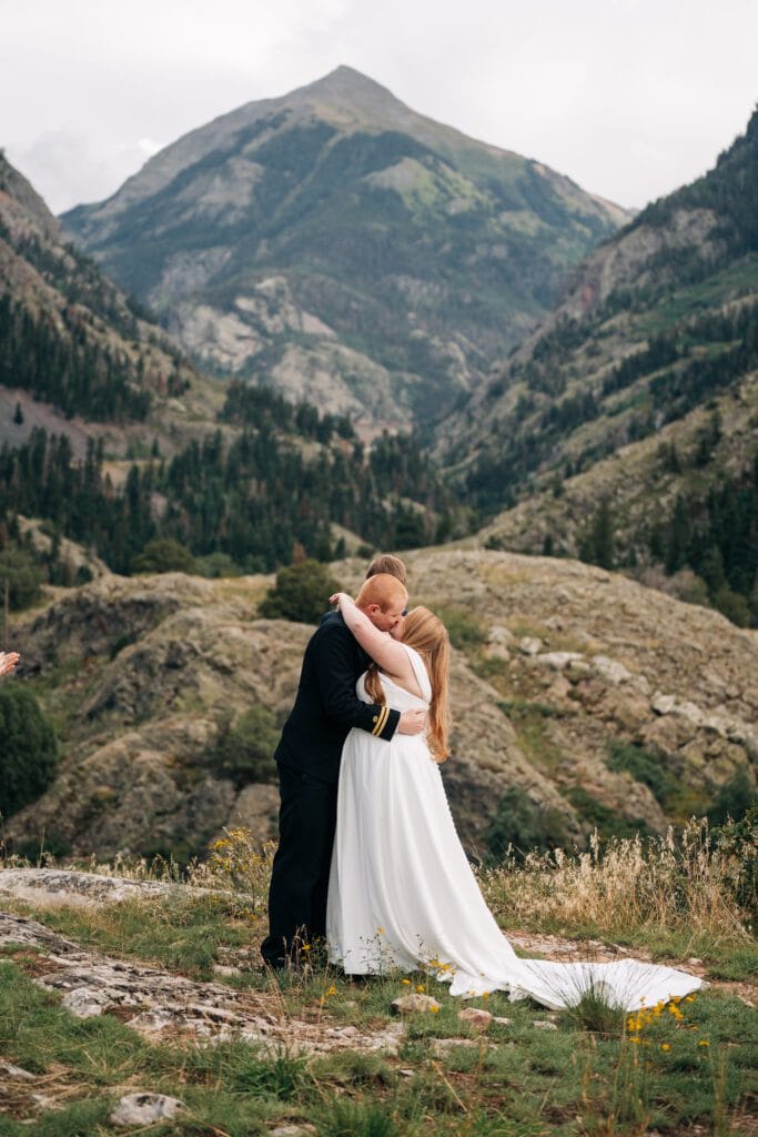 A bride and groom embrace and kiss outdoors, surrounded by rocky hills and tall mountains, with greenery and a cloudy sky in the background.