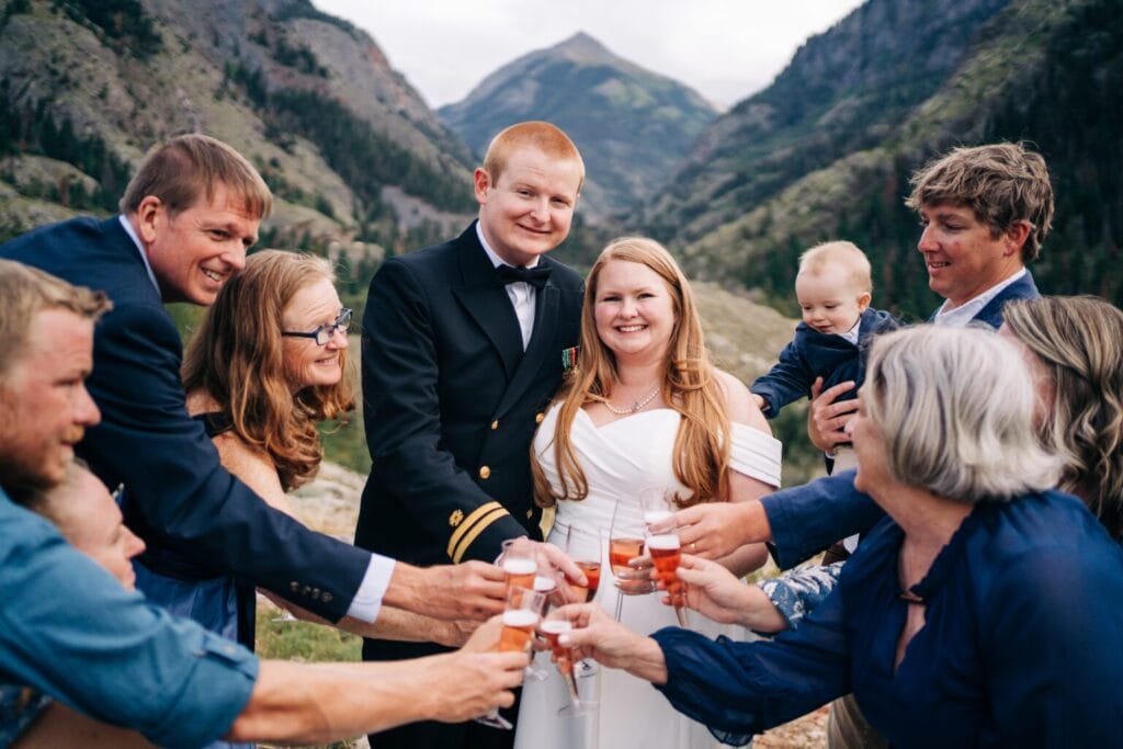 A bride and groom, surrounded by family and friends, smile and raise glasses in a toast outdoors in a mountainous landscape. Everyone appears happy and celebratory, with a baby also present in the group.