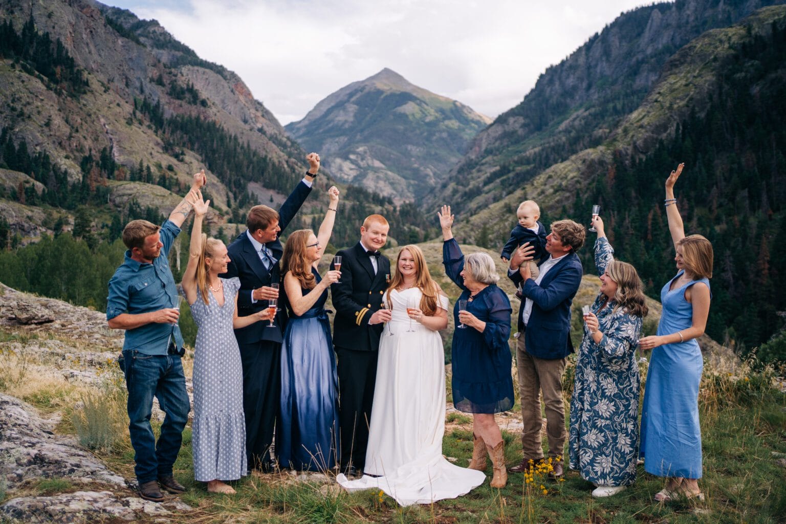 A wedding party celebrates outdoors in a mountain valley. The bride and groom stand in the center, surrounded by smiling family and friends with raised arms, with lush mountains in the background.
