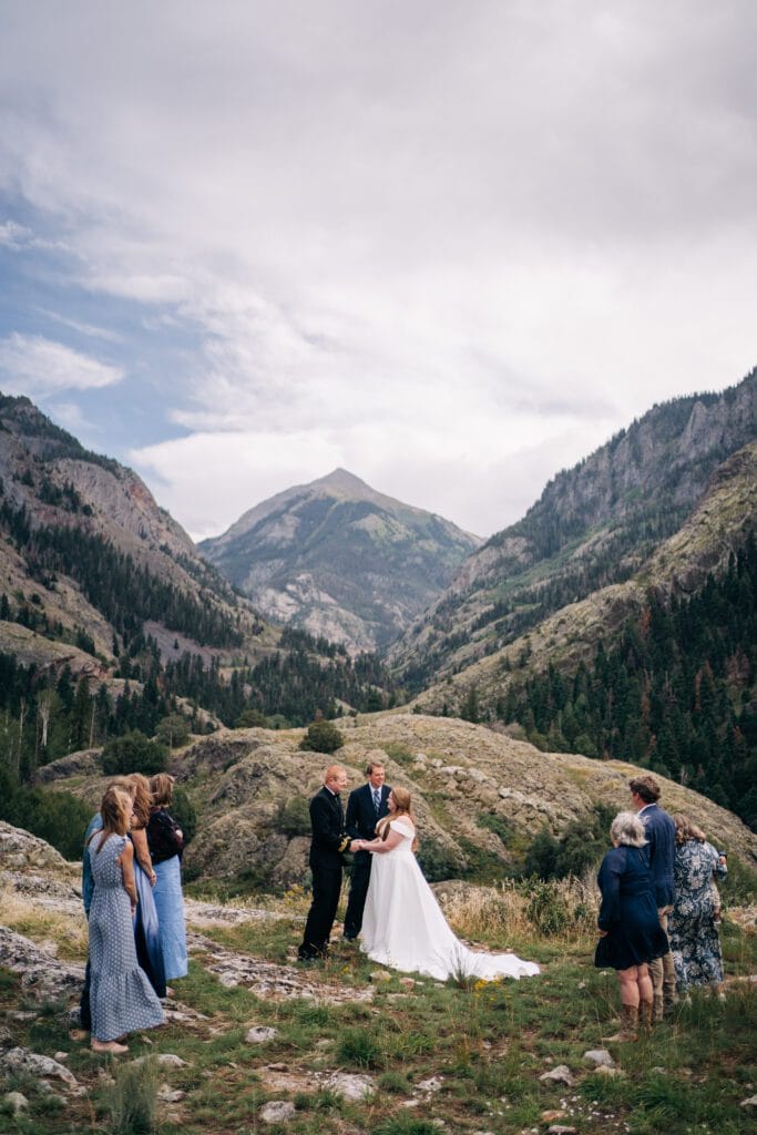 A bride and groom stand holding hands during an outdoor wedding ceremony surrounded by guests in a scenic mountain landscape with lush green hills and a cloudy sky.