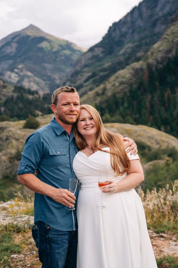 A smiling couple stands outdoors in front of mountains, with the woman in a white dress and the man in a blue shirt; both are holding glasses and embracing each other.