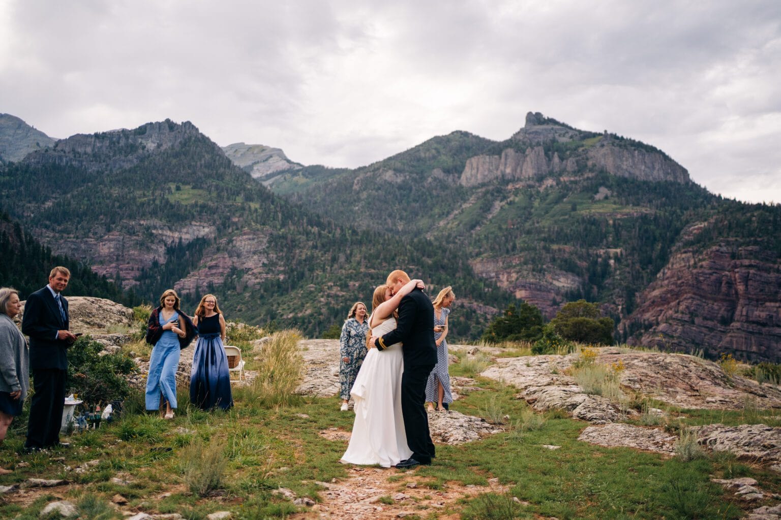 A bride and groom embrace and kiss outdoors on a rocky, grassy area with mountains in the background, while a small group of guests looks on and smiles under a cloudy sky.
