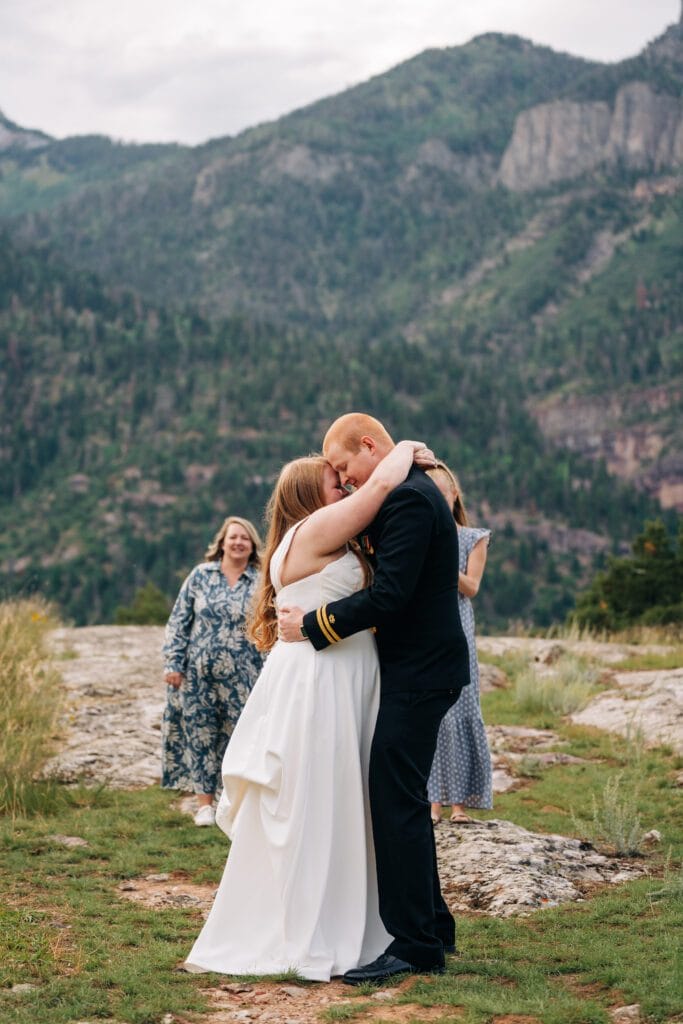 A bride and groom embrace and kiss outdoors on a rocky landscape with green mountains in the background. Two women stand behind them, smiling and watching the couple.