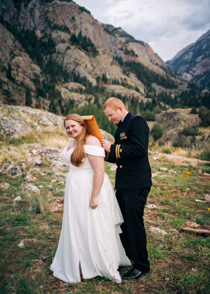A smiling bride in a white dress stands outdoors as a man in a navy military uniform attaches a large yellow envelope to her back, with mountains and greenery in the background.