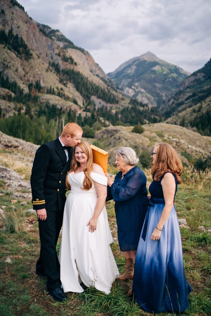 A bride in a white dress stands in a mountain landscape, smiling as a man in a naval uniform kisses her head. Two women in blue dresses help adjust her dress outdoors, surrounded by greenery and rocky hills.