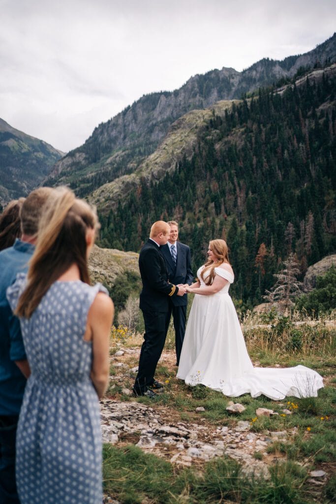 A bride and groom stand outdoors holding hands during a wedding ceremony in front of a mountain landscape, with a few guests watching from the side. The bride wears a white dress and the groom a black suit.