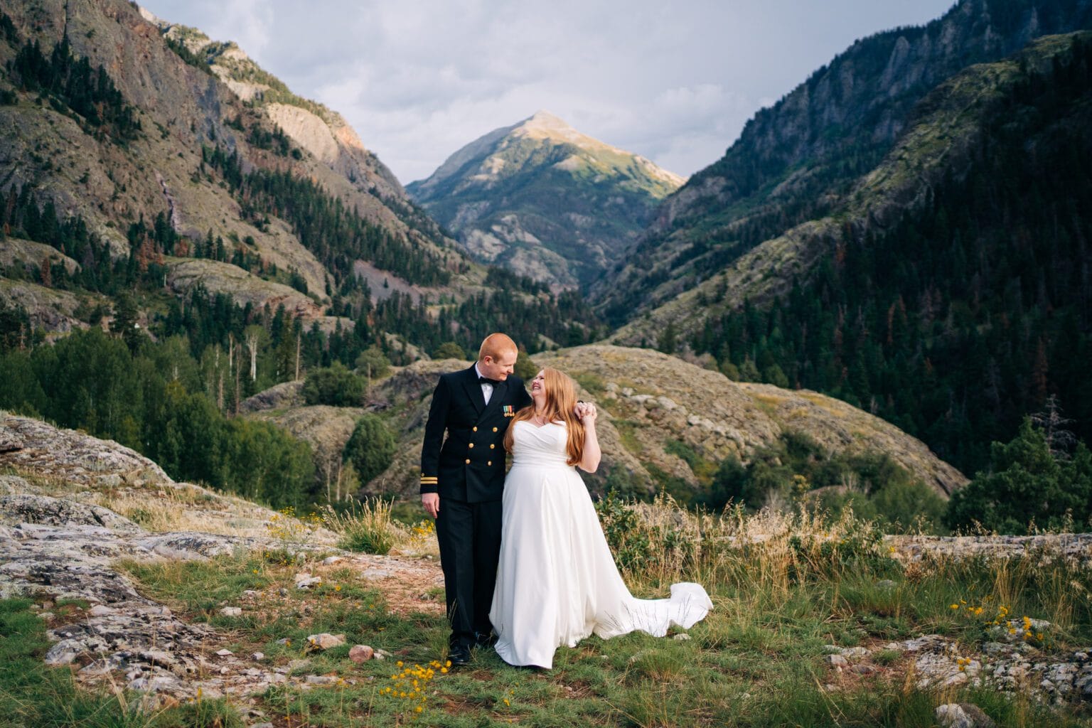 A bride in a white dress and a groom in a navy uniform stand together on rocky terrain, surrounded by green mountains and valleys, with a distant snow-capped peak under a cloudy sky.