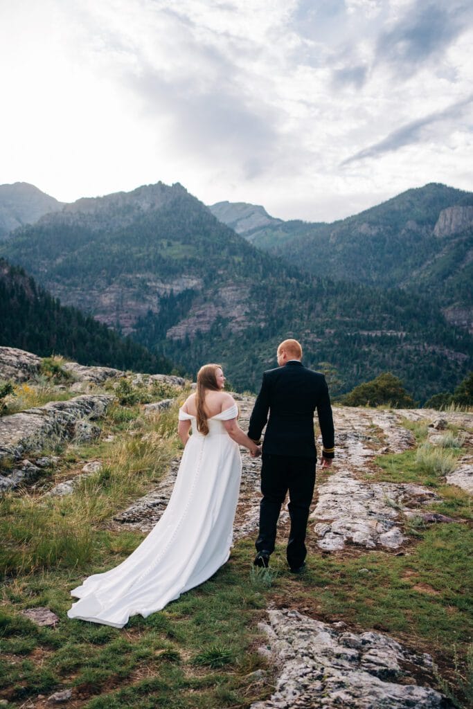 A bride in a white dress and a groom in a black suit walk hand in hand along a rocky path, surrounded by lush mountains and a cloudy sky.