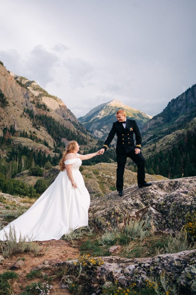 A bride in a white dress and a groom in a black suit stand on rocky terrain, holding hands, surrounded by green mountains and a dramatic sky in the background.