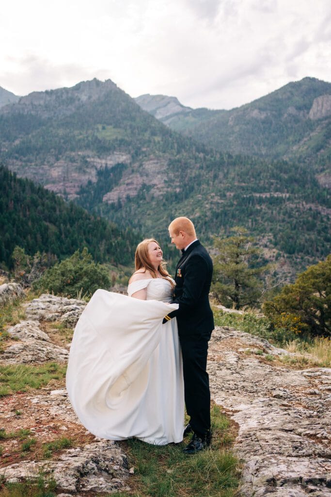 A bride and groom stand on a rocky mountain overlook surrounded by pine trees. The bride’s white dress billows as they smile at each other, with scenic forested mountains and a cloudy sky in the background.