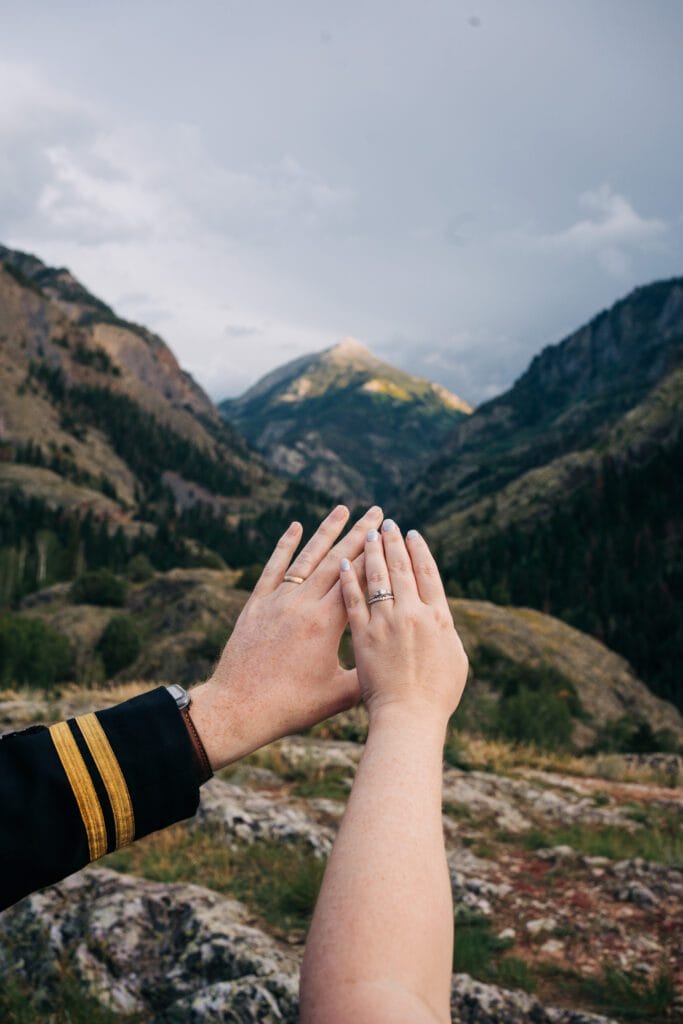 Two hands with wedding rings held together in front of a scenic mountain landscape, with one person wearing a dark sleeve with a gold stripe.