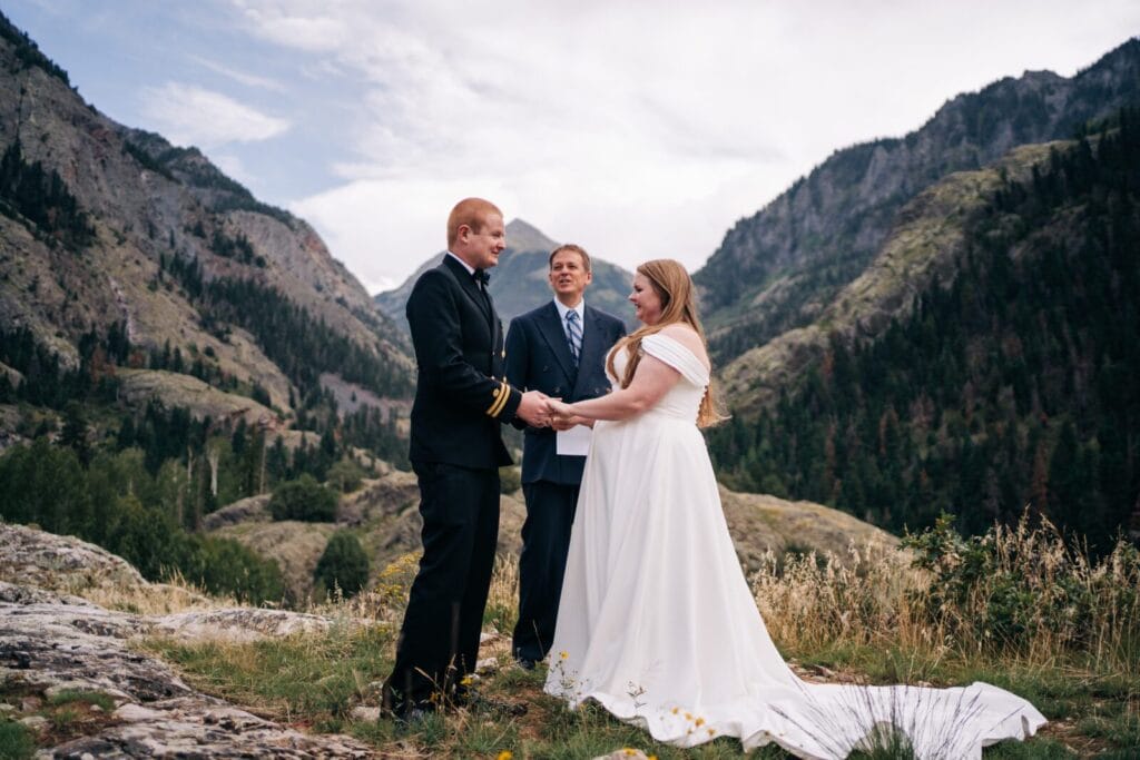 A bride and groom stand holding hands during an outdoor wedding ceremony with an officiant, surrounded by green mountains and a cloudy sky. The bride wears a white dress, and the groom wears a dark suit with gold trim.
