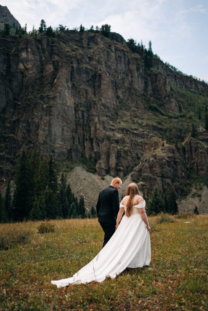 A bride in a flowing white dress and a groom in a black suit walk hand in hand through a grassy field, with tall cliffs and pine trees rising in the background.