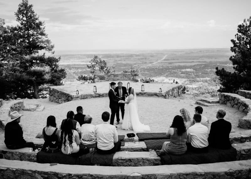 A couple stands with an officiant, exchanging vows at an outdoor stone amphitheater overlooking a vast landscape, while a small group of seated guests watches. The scene is in black and white.