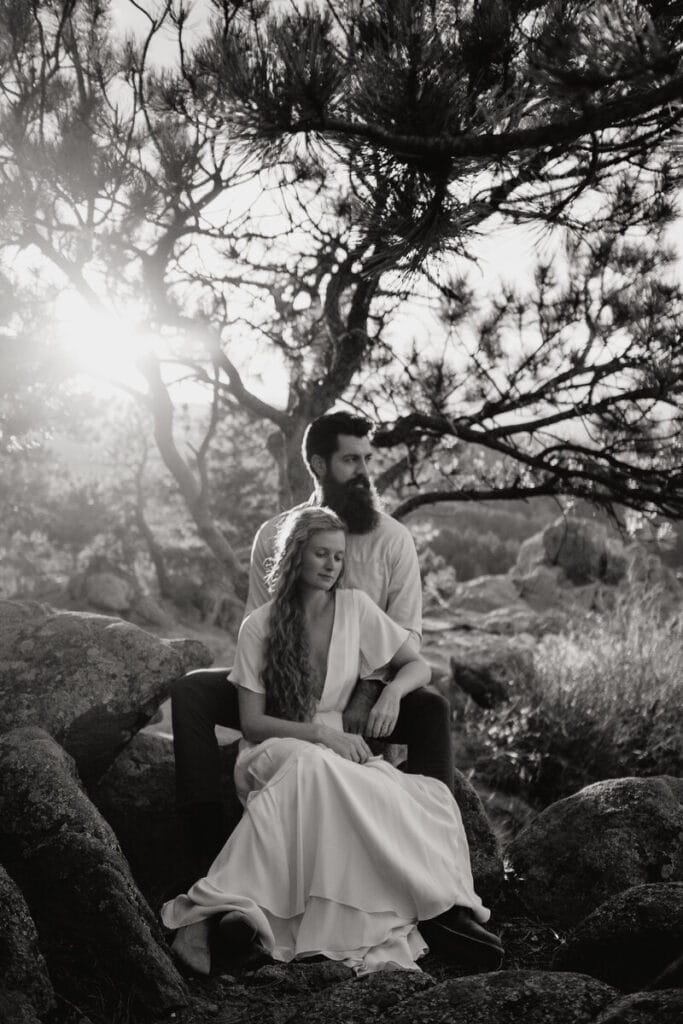 A couple sits on rocks beneath a tree, with sunlight filtering through branches. The woman in a white dress sits in front, eyes closed, while the bearded man sits behind, holding her gently. The scene is peaceful and intimate.