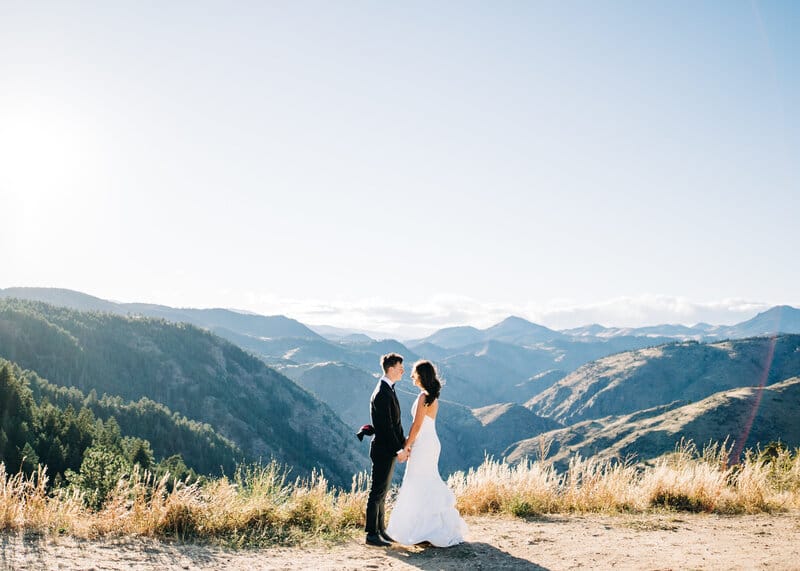 A bride and groom stand hand in hand on a grassy hilltop, facing each other, with mountains and a clear blue sky in the background.