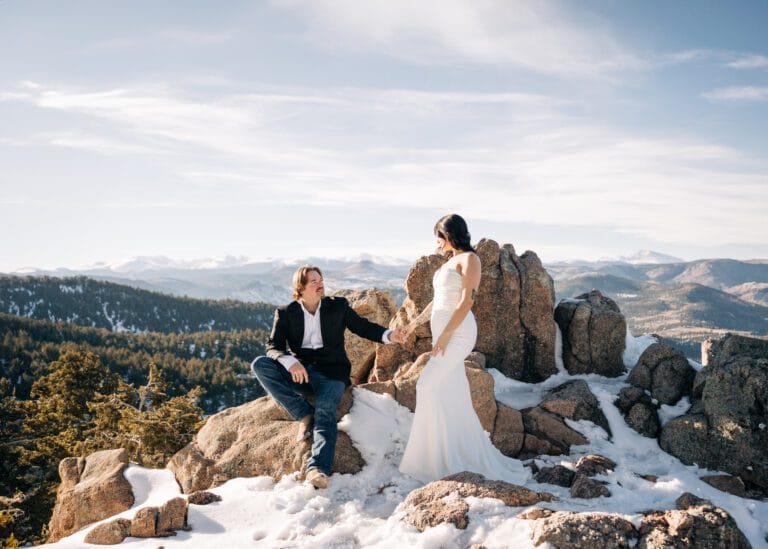 groom sitting on rock and offering his hand to his wife during their boulder elopement at Lost Gulch Overlook